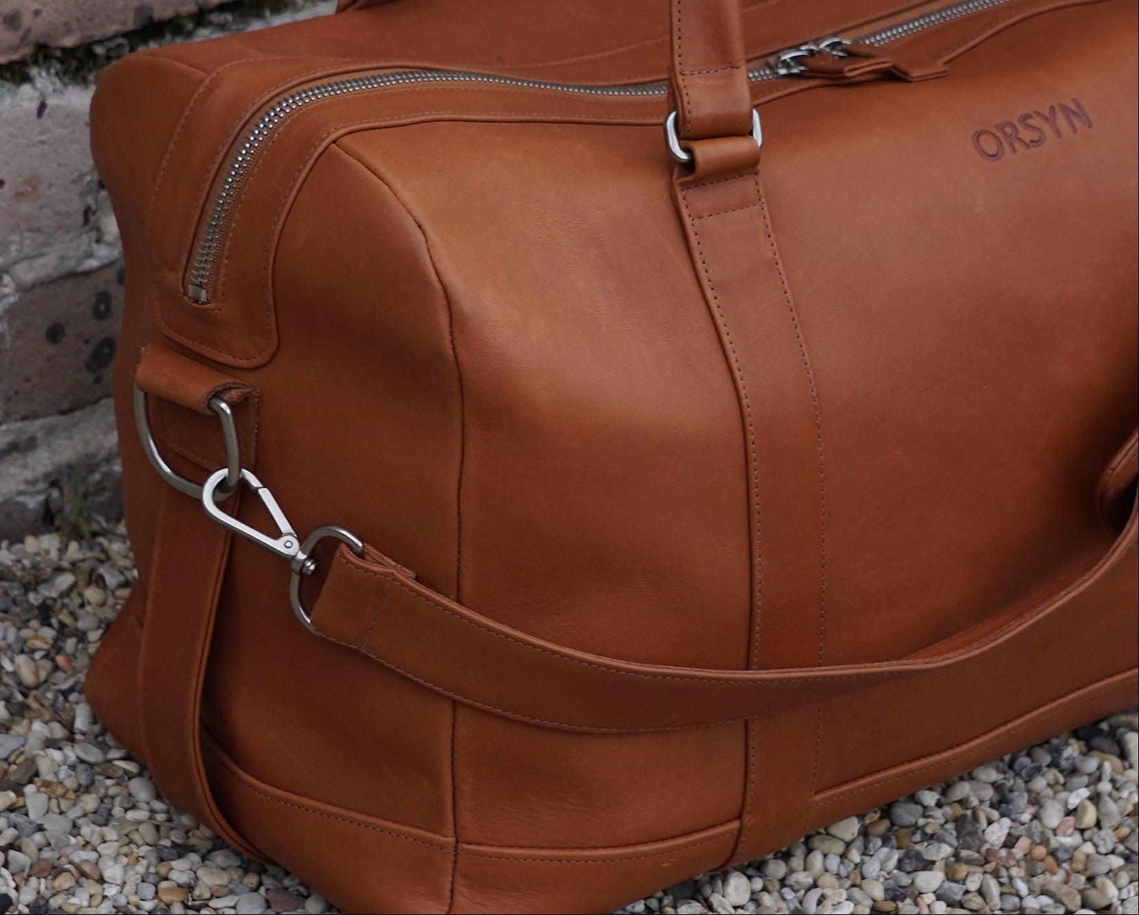 Brown leather duffel bag with 'ORSYN' branding on a stone surface.